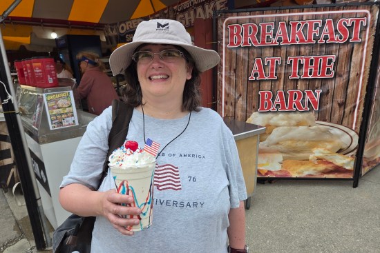Anne smiling and holding a red, white and blue shake in front of a large sign reading "Breakfast at the Barn".