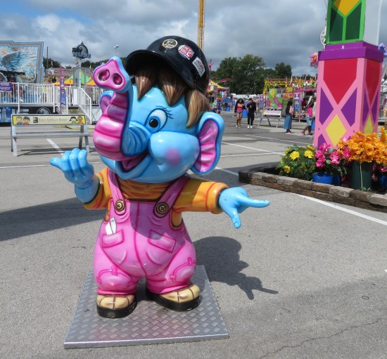 Statue of elephant boy in purple overalls and black baseball helmet, in front of carnival rides.