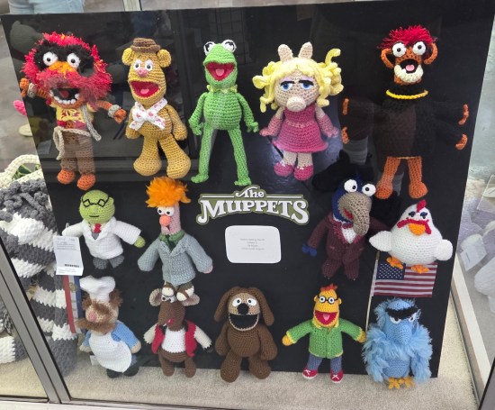 14 crocheted Muppet dolls hanging on a black backing board.