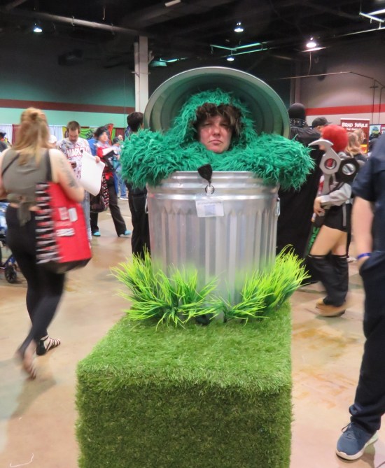 Oscar the Grouch cosplayer, green fuzzy suit inside a trash can atop a mobile platform, frowning hard.