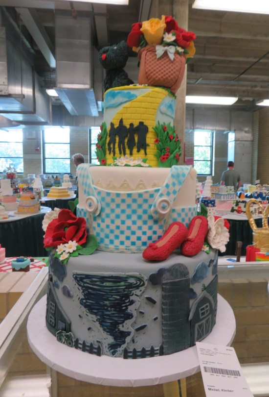 Three-layer cake with layers of black-and-white tornado, Dorothy's blue farmgirl dress, and the Yellow Brick Road. A Toto doll and Dorothy's basket are on top.