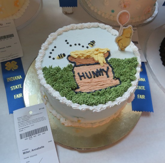 Single-layer cake with honeypot drawing in icing, and a little cardboard Pooh standing on it.