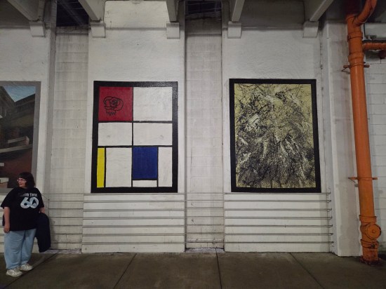 Replicas of Piet Mondrian and Jackson Pollock paintings on a gray concrete wall. Anne stands at left.