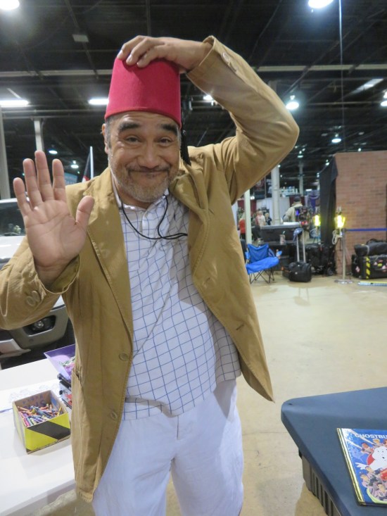 Sallah cosplayer, holding his red fez on with one hand and waving hi with the other.