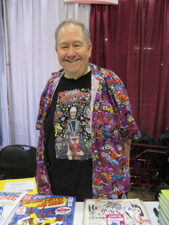 Scott Shaw at his convention table, wearing a purple Svengoolie shirt over a black Svengoolie T-shirt.