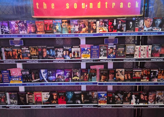Four wall racks filled with movies on videotape. Each shelf is lined with reminders to "Be Kind Rewind".