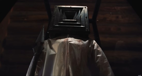 Murderer wearing white jumpsuit and black box on head stands with spear at attention like a soldier