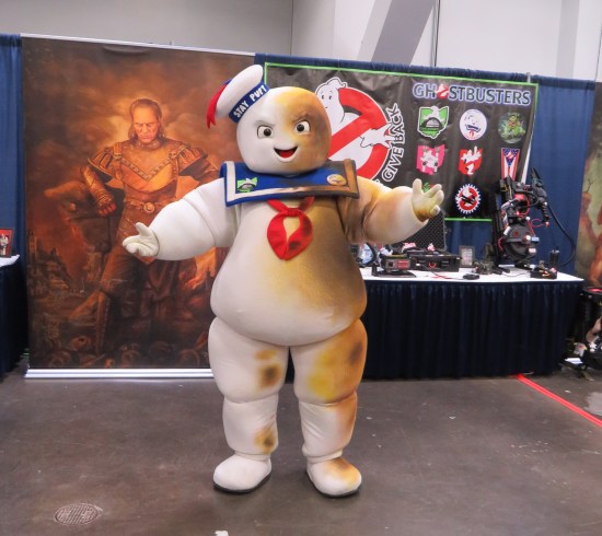 Mr. Stay-Puft cosplayer but with much singeing on their left side, in front of a Ghostbusters cosplay club booth.