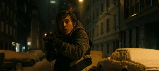 Scared young white guy on a wintry city street vaguely points an assault rifle.