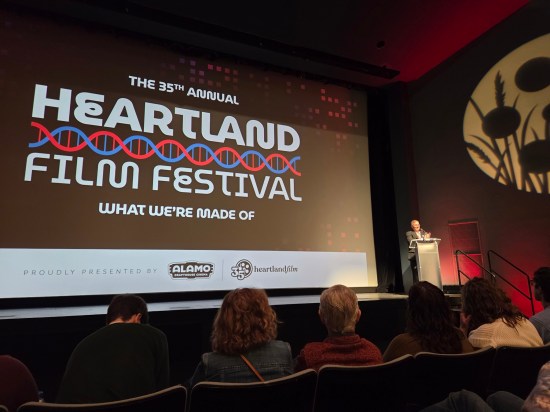Theater screen with Heartland logo on it. Next to it, a man speaks at a podium.