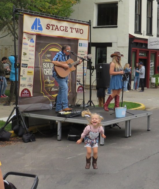 Young lady and older gentleman do country music on an outdoor stage while a toddler bounces up and down in front.