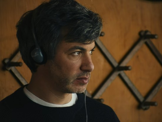 Nathan Fielder a wood-paneled kitchen with headphones and weird stare.