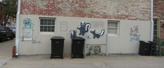 Back of a building lined with trash cans. Painted above them are pics of skunks, raccoons, and Oscar the Grouch.
